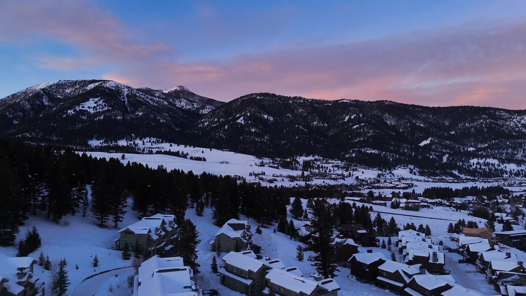 Snow-covered mountain neighborhood with pines and rooftops at dusk